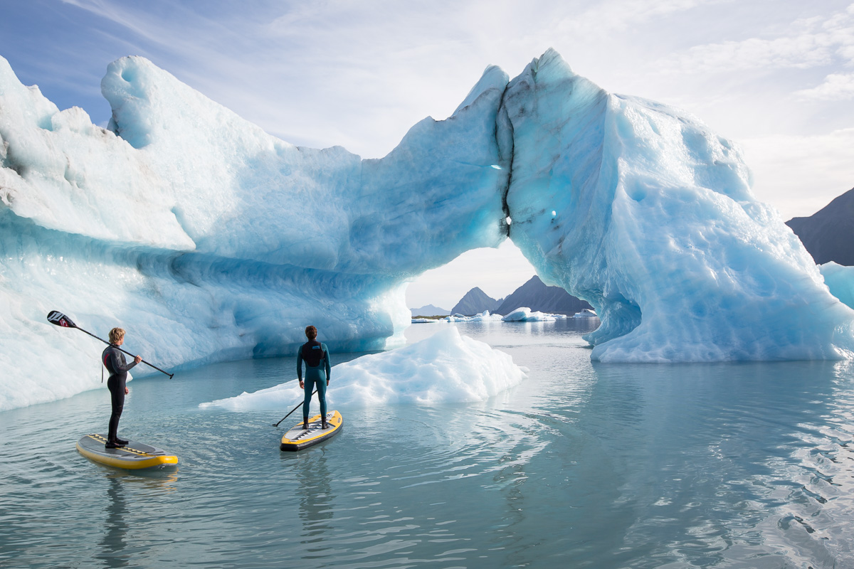 Kevin Langeree and Kai Lenny stand up paddle around a large iceberg with an arch formed in it in a glacier lake in Southcentral Alaska.